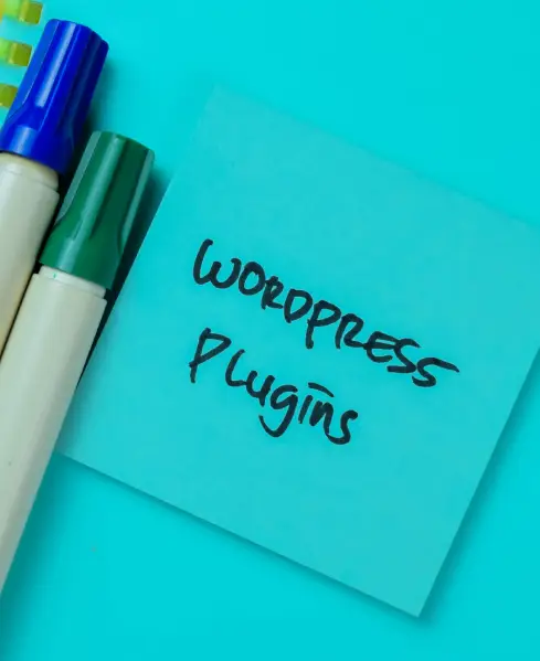 A blue sticky note with the handwritten text "WORDPRESS Plugins" in black marker, sitting next to a blue and a green marker on a bright blue surface.